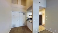 This single-floor residential space is thoughtfully arranged to maximize functionality and flow. The living area is anchored by plush carpeted flooring and benefits from impressively tall ceilings that lend an airy, open feel to the room. A set of double sliding-door closets provides generous built-in storage, making the space as practical as it is comfortable.

The kitchen is efficiently designed with a pass-through counter layout, offering dark countertops that contrast neatly with white upper cabinetry. Black appliances, including a range and a refrigerator, are integrated into the kitchen, complemented by an overhead ventilation hood. A sink is conveniently positioned along the counter, and the kitchen's open orientation keeps it visually connected to the surrounding living space.

A central dividing wall subtly separates the living area from the hallway corridor, which transitions to hard-surface flooring — providing a natural delineation between spaces. The hallway leads further into the unit, hinting at additional rooms beyond. Neutral wall tones throughout create a clean, versatile backdrop. HVAC vents are present in multiple areas, ensuring climate comfort. Overall, the space presents a practical and well-organized single-level layout suited for everyday living.