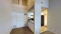 This single-floor residential space is thoughtfully arranged to maximize functionality and flow. The living area is anchored by plush carpeted flooring and benefits from impressively tall ceilings that lend an airy, open feel to the room. A set of double sliding-door closets provides generous built-in storage, making the space as practical as it is comfortable.

The kitchen is efficiently designed with a pass-through counter layout, offering dark countertops that contrast neatly with white upper cabinetry. Black appliances, including a range and a refrigerator, are integrated into the kitchen, complemented by an overhead ventilation hood. A sink is conveniently positioned along the counter, and the kitchen's open orientation keeps it visually connected to the surrounding living space.

A central dividing wall subtly separates the living area from the hallway corridor, which transitions to hard-surface flooring — providing a natural delineation between spaces. The hallway leads further into the unit, hinting at additional rooms beyond. Neutral wall tones throughout create a clean, versatile backdrop. HVAC vents are present in multiple areas, ensuring climate comfort. Overall, the space presents a practical and well-organized single-level layout suited for everyday living.