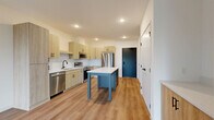 Located at 11816 Wayzata Blvd in Minnetonka, MN, this modern one-bedroom, one-bathroom unit offers a well-designed single-floor layout with contemporary finishes throughout. The open-concept kitchen and living area serves as the heart of the space, featuring light wood cabinetry, a white subway tile backsplash, stainless steel appliances including a refrigerator, dishwasher, and range, and a central kitchen island with a crisp white countertop. Recessed lighting and durable wood-style flooring run throughout the main living area, which is bathed in natural light from large floor-to-ceiling windows and a sliding glass door that opens to a private balcony with views of surrounding greenery. The bedroom is a comfortable, carpeted retreat equipped with a ceiling fan, a large black-framed window overlooking lush outdoor scenery, and ample closet access. The bathroom features a warm wood vanity with black hardware, a white countertop, a tiled walk-in shower, and wood-style flooring that ties cohesively to the rest of the unit. Additional spaces include a laundry room, utility room, and multiple closets, providing generous storage throughout. The overall aesthetic is clean, modern, and bright.