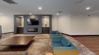 This single-floor space is thoughtfully designed to blend functional amenities with contemporary style. The kitchen is a standout feature, outfitted with dark espresso cabinetry, light marble-veined quartz countertops, and a full suite of stainless steel appliances, including a refrigerator, dishwasher, microwave, and a cooktop with a stainless steel range hood. A central island provides additional prep and gathering space, while rich hardwood-style flooring ties the area together with warmth and durability.

The open layout transitions seamlessly into a spacious lounge area, where a slate-blue accent wall with natural stone detailing serves as a dramatic backdrop for a wall-mounted television and a sleek linear fireplace. Built-in cabinetry flanks the feature wall, adding both function and visual balance. Seating throughout includes teal plaid sofas and modern swivel lounge chairs arranged around low-profile tables, creating comfortable conversation areas.

Large black-framed windows along the perimeter flood the space with natural light and offer views of an outdoor courtyard. Patterned carpeting in the lounge zone contrasts with the hardwood flooring, subtly delineating areas within the open plan. Recessed lighting throughout ensures a well-lit, inviting atmosphere.