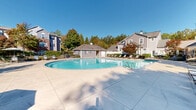 Located at 5500 Oakley Industrial Blvd in South Fulton, GA, this outdoor amenity area centers around a generously sized swimming pool that serves as the focal point of the space. The pool features clearly marked lap lanes for fitness swimming alongside a broader open swimming area, accommodating both recreational and exercise use. Wide concrete pool decking surrounds the water on all sides, providing abundant space for lounging and circulation.

A dedicated pool house structure anchors one end of the complex, finished with siding and a standing-seam metal roof that complements the architectural style of the surrounding multi-story residential buildings. The pool house appears to offer utility and support functions for the pool area. Numerous lounge chairs and chaise lounges are arranged throughout the deck, offering residents ample seating options.

The perimeter of the pool area is framed by lush, mature trees displaying vibrant autumn foliage, lending a natural, park-like ambiance to the setting. Decorative lamp posts are positioned throughout, ensuring the space is well-lit during evening hours. A separate, smaller wading or spa pool is also visible along one side of the facility. The entire area benefits from abundant natural sunlight under open skies, creating a bright and inviting outdoor retreat for residents of the surrounding community.