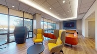 Located at 4400 E West Hwy in Bethesda, MD, this single-floor commercial lounge space delivers a dynamic, contemporary environment designed to foster comfort and connectivity. The open layout is anchored by expansive floor-to-ceiling windows that flood the interior with natural light and offer clear views of the surrounding streetscape, creating an airy, welcoming atmosphere throughout.

The seating area is thoughtfully curated with a bold, modern aesthetic. High-back wing chairs in yellow and gray tones are paired with vibrant orange accent seating, providing a variety of informal gathering spots. Hexagonal pod-style chairs add additional semi-private seating options, ideal for focused conversations or quiet reflection. Warm hardwood-style flooring runs throughout, contrasting elegantly with the sleek tile ceiling system fitted with recessed and cove lighting.

A textured deep-blue accent wall serves as a striking design element, complemented by a wall-mounted flat-screen display and warm wood cabinetry — forming a polished reception or media zone. Adjacent to the main lounge, a kitchenette alcove features dark mosaic tile backsplash, pendant lighting, and built-in appliances, adding functional amenity to the space.

The overall ambiance is energetic yet inviting, balancing professional utility with vibrant design — well-suited for a client-facing lobby, collaborative workspace, or community lounge.