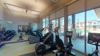 This fitness center is a thoughtfully designed, single-floor facility that offers a comprehensive workout environment for users of all fitness levels. The space is divided into distinct functional zones, each tailored to a specific type of exercise. The cardio area is lined with a generous row of treadmills, ellipticals, and stationary bikes — including Peloton-style cycling bikes — all positioned along expansive floor-to-ceiling windows that flood the space with natural light and provide open views of an outdoor courtyard and surrounding community. Roller shades offer flexible light control throughout the day. The strength and functional training area features a rowing machine, cable and weight machines, and a rack stocked with medicine balls, supporting a wide range of workout routines. An open stretch and movement zone provides ample floor space, accented by contemporary artwork on the walls and a sleek built-in counter with storage. The interior design is clean and modern, with two-tone flooring in soft yellow-green and deep charcoal, elevated tray ceilings with ceiling fans, and warm wood slat wall panels that add a sophisticated architectural detail. Wall-mounted televisions and mirrored walls further enhance the functional and airy atmosphere of this well-appointed fitness facility.