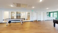 Located at 99 Van Block Ave in Hartford, Connecticut, this single-floor community space offers a thoughtfully designed kitchen and living area that combines functionality with a lively, modern aesthetic. The kitchen is outfitted with white shaker cabinetry, granite countertops, stainless steel appliances including a refrigerator, dishwasher, and microwave, and a striking green ceramic tile backsplash that adds a bold design accent. A pendant light and recessed lighting illuminate the space, while a large granite-topped island provides additional prep and gathering space. Light wood-toned flooring flows seamlessly throughout the entire area, contributing to a warm and cohesive feel. The living area is defined by a deep navy blue accent wall, comfortable tan leather sofas arranged around a central coffee table, and a wall-mounted flat-screen television. Large windows along the green-accented walls flood the space with natural light and offer views of the surrounding Hartford neighborhood. A glass partition separates a game zone featuring a shuffleboard table and foosball table, adding a recreational dimension to the space. The open layout, vibrant color palette, and variety of seating and activity areas make this a welcoming and versatile community gathering space.