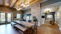 Located at 24500 Wildwood Park Rd in Richmond, TX, this single-floor amenity space is designed to blend comfort and functionality in a cohesive, stylish environment. The living room serves as a welcoming lounge area, featuring warm hardwood flooring, striking gold drum pendant lights, and a plush mustard-yellow sofa anchored by a patterned area rug. A built-in teal shelving unit with decorative accents and a wall-mounted TV adds both character and practicality, while large windows invite natural light throughout. Framed abstract artwork in gold finishes adorns the sage-green walls, contributing to a refined yet approachable atmosphere. The primary dining area showcases exposed wood beam ceilings that lend a rustic-modern charm, centered around a substantial farmhouse-style dining table with mixed seating. Adjacent to it, a dedicated conference-style dining room offers a more formal setting, complete with a rich hardwood table, upholstered rolling chairs, a wall-mounted display screen, and an elegant geometric gold chandelier beneath a skylit ceiling. The kitchen is outfitted with deep navy cabinetry, light stone countertops, glass-front upper cabinets, and a tiled backsplash, offering generous prep and serving space alongside a window overlooking the outdoors. Together, these spaces create a versatile, well-appointed environment suited for gathering, dining, and collaboration.