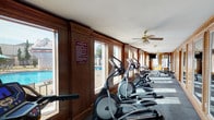 This fitness center is a single-floor facility that combines classic architectural character with a comprehensive selection of exercise equipment. The interior is defined by rich, dark wood panel walls framed with detailed molding, lending the space a warm and distinguished aesthetic that sets it apart from a typical gym environment. Large windows line one entire wall, flooding the room with abundant natural light and offering direct views of an outdoor swimming pool and surrounding patio area, creating a bright and motivating atmosphere throughout.

The facility is generously outfitted with a wide variety of cardio equipment, including treadmills, elliptical trainers, and stationary and spinning bikes, arranged in a long, open layout that promotes easy movement between stations. A dedicated free weights area features a full rack of dumbbells alongside a rowing machine, providing options for strength and resistance training. A multi-use cable and weight machine is positioned at one end of the room, rounding out the strength training capabilities. Protective rubber flooring tiles cover the workout areas, ensuring safety and comfort during exercise.

A wall-mounted television is available for entertainment during workouts, and a ceiling fan helps maintain airflow throughout the space. A posted fitness center rules sign reflects the managed and community-oriented nature of the facility. Entry is provided through a traditional paneled door with an arched window detail, maintaining the cohesive classic design of the space.