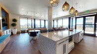Located at 2420 Alcott Street in Denver's vibrant LoHi neighborhood, this sophisticated amenity lounge offers an elevated experience with panoramic views of the Denver skyline. Floor-to-ceiling windows wrap the space, flooding the interior with natural light and framing stunning city and mountain vistas from every angle.

The open-plan layout is anchored by a large granite-topped kitchen island with bar seating, pendant lighting in smoked glass, and white cabinetry. The full-length kitchen features a striking blue arabesque tile backsplash, granite countertops, stainless steel appliances, and ample counter space — ideal for catering and entertaining.

A dedicated games and lounge area includes a billiards table, a large wall-mounted flat-screen TV set against a bold blue accent wall, and comfortable leather seating arranged around a textured coffee table. A separate seating nook with floor-to-ceiling corner windows provides an intimate retreat with direct access to the outdoor terrace, where lounge chairs and a pool are visible.

Rich hardwood flooring runs throughout, complemented by contemporary track lighting and circular overhead fixtures. The space also features a dining area with modern seating and sculptural tables. Together, these elements create a dynamic, well-appointed communal space designed for socializing, relaxation, and enjoying Denver's impressive skyline.