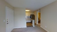 This single-floor residential unit offers a practical and well-organized open-concept layout designed for comfortable everyday living. The living area is finished with plush wall-to-wall carpeting, creating a warm and inviting atmosphere, while the kitchen and adjoining spaces transition to durable hardwood-style flooring for easy maintenance.

The kitchen is a functional centerpiece of the home, featuring white cabinetry, granite-style countertops, and a decorative tile backsplash with a subtle mosaic pattern. A stainless steel refrigerator anchors the space, and a peninsula-style breakfast bar extends outward, offering additional prep space and a natural divider between the kitchen and living area. Overhead flush-mount lighting keeps the space bright and well-illuminated.

A hallway visible from the main living area leads to the additional rooms, including what appears to be a bedroom or secondary space with its own cabinetry or built-in storage, maintaining a consistent design aesthetic throughout. The unit features clean white trim, neutral beige walls, and a textured ceiling, all contributing to a cohesive and understated interior palette. The overall layout is efficient and thoughtfully arranged, making the most of the available space on a single level.