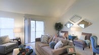 This two-floor residence offers a thoughtfully designed living environment with a warm and inviting ambiance throughout. The main living area immediately draws attention with its vaulted ceilings, which lend an airy sense of openness to the room. A large sliding glass door floods the space with natural light and provides direct access to a private balcony, where views of the surrounding outdoors can be enjoyed. The living room is generously proportioned, offering ample space for comfortable seating arrangements and furnishings. Soft, neutral wall tones complement the carpeted flooring, creating a relaxed and cozy atmosphere. Decorative touches, including wall-mounted mirrors and accent furniture, add character to the space. The layout across two floors provides a natural separation between living and private areas, offering both functionality and comfort. The overall design balances practicality with a light, casual aesthetic, making the space well-suited for everyday living. The combination of vaulted ceilings, abundant natural light, and outdoor access through the balcony are among the most standout architectural features this residence has to offer.