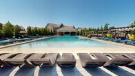 Located at 3800 SW Richsmith Rd in Bentonville, Arkansas, this two-floor community amenity complex is anchored by a resort-style outdoor pool area designed for relaxation and recreation. The centerpiece is a generously sized swimming pool with crystal-clear water, flanked on all sides by an expansive sun deck furnished with rows of wicker chaise lounges outfitted with vibrant blue cushions. A separate lap pool, framed by decorative brick columns topped with bowl-shaped fire features, provides a dedicated space for fitness-focused swimmers. The pool area is bordered by mature evergreen trees that offer privacy and a natural backdrop. Shaded seating zones featuring wicker sectional sofas with blue cushions provide comfortable gathering spaces throughout the deck. A covered pergola along one side extends the outdoor living experience with additional lounge seating. The adjacent brick clubhouse serves as the architectural focal point, boasting a classic pitched roofline, a prominent exterior chimney, and an expansive wall of floor-to-ceiling glass that floods the interior with natural light. Lush potted plants and colorful floral arrangements add vibrancy throughout the space, while well-maintained green turf areas complement the hardscaped pool surround, creating a polished and resort-caliber outdoor environment.