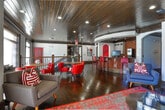Located at 100 Perimeter Trace E in Dunwoody, Georgia, this single-floor communal space is a well-appointed clubroom that blends upscale design with a welcoming social atmosphere. Rich, dark hardwood floors run throughout, complemented by matching dark wood-planked ceilings with recessed lighting that create a warm, intimate ambiance.

The centerpiece of the space is a curved bar adorned with a mosaic tile facade and dark granite countertop, accompanied by bold red bar stools. Behind the bar, a full kitchen area features white cabinetry, tile backsplash, and modern appliances, making it well-suited for hosting and entertaining.

Adjacent to the bar, a lounge area offers comfortable gray mid-century-style seating arranged around a coffee table atop a vibrant patterned area rug. A wall-mounted flat-screen television anchors the lounge, while large windows with blinds allow natural light to filter in. Curated artwork, including large abstract and figurative pieces, adds a gallery-like quality throughout.

A separate dining area provides round tables paired with tufted red leather chairs, creating an ideal setting for casual gatherings. Iron stair railings frame an open stairwell, adding an architectural detail that enhances the overall sophisticated aesthetic of this stylishly designed community space.
