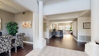 The Jovanna Villas community clubhouse is a thoughtfully designed single-floor facility that serves as a central gathering hub for residents. The grand entrance features elegant white columns, large-format tile flooring transitioning to rich hardwood, and soaring ceiling heights that create an immediately impressive first impression. Colorful artwork adorns the neutral gray walls, setting a welcoming tone throughout.

The interior flows naturally from the entry lobby into a series of well-appointed common areas. A comfortable lounge space features plush gray sofas, a patterned area rug, and windows dressed with wood-slatted blinds that allow ample natural light while offering views of the surrounding landscape. A sun-drenched sitting room at the rear of the building boasts expansive windows and glass doors that open directly to the outdoor pool and patio area, seamlessly blending indoor and outdoor living.

A dedicated dining room with dark hardwood floors accommodates a large table and is accented by a tasteful abstract art piece. The kitchen is equipped with white cabinetry, dark countertops, and stainless steel appliances, ideal for community events and gatherings.

The Jovanna Fitness Gym is a well-equipped exercise space featuring cardio machines, a multi-station weight machine, a stair climber, ceiling fans, wall-mounted televisions, and floor-to-ceiling mirrors that amplify the sense of space.