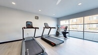 Located at 99 Glenview in Ontario, Ohio, this single-floor fitness center offers a clean, modern environment designed to support a well-rounded workout routine. The space is finished with light hardwood-style flooring throughout, providing a bright and contemporary aesthetic that complements the neutral gray and white wall palette.

The cardio area is well-equipped with multiple Star Trac treadmills and a recumbent exercise bike, all arranged to take advantage of the large floor-to-ceiling windows along one wall. These expansive windows allow generous natural light to fill the space and offer views of the surrounding property. A ceiling fan with integrated lighting and recessed ceiling lights ensure the room remains comfortable and well-lit during any hour.

On the strength training side, a multi-function cable machine provides a variety of resistance-based exercises, with an instructional exercise chart mounted nearby for easy reference. Sanitizing wipes and a waste bin are conveniently stationed in the space, reflecting an emphasis on cleanliness and user convenience.

A wall-mounted mini-split HVAC unit provides independent climate control, ensuring year-round comfort. An adjacent restroom is accessible directly from the fitness area. The overall design is minimalist and functional, offering a welcoming environment for residents seeking a convenient, on-site fitness facility.