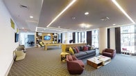 This contemporary clubroom is thoughtfully designed to offer a dynamic and inviting social environment. The space is organized into distinct yet open zones, allowing residents or guests to gather, relax, and entertain with ease. A modern lounge area anchors the room, furnished with plush gray sofas and curved, sculptural armchairs in warm mauve tones, accented by colorful throw pillows in yellow, red, and pink for a lively pop of color. Sleek, low-profile coffee tables complement the seating arrangement and add to the refined aesthetic. Adjacent to the lounge, a full-size billiards table provides a central entertainment focal point. Along the back wall, a built-in shelving unit with warm wood accents houses a large flat-screen television, display shelves, and additional seating nooks, creating a dedicated media and activity zone. Recessed lighting and continuous LED cove lighting along the ceiling provide a warm, layered illumination throughout. Large floor-to-ceiling windows along one side of the room flood the space with natural light and offer views of the surrounding exterior. The patterned carpet, neutral wall tones, and carefully curated furnishings come together to create a space that is both functional and visually sophisticated.