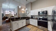 Located at 1278 W Ninth Street in Cleveland's vibrant Ohio City neighborhood, this single-floor loft seamlessly combines industrial heritage with contemporary design. The open-concept living and kitchen area is anchored by soaring ceilings, hardwood floors, and striking exposed brick walls framed by large industrial-style windows that flood the space with natural light. The kitchen is well-appointed with white shaker cabinetry, dark granite countertops, a classic subway tile backsplash, stainless steel appliances, and a breakfast bar with seating — ideal for casual dining and entertaining. Pendant lighting adds a warm, modern touch overhead. The living area features an inviting layout with ample seating and direct sightlines to the kitchen. Two bedrooms offer comfortable retreats, each with carpeted floors and interior clerestory windows that borrow light from the adjoining spaces. Both bathrooms are finished with granite-topped vanities, tile or wood-look flooring, and tub-shower combinations. Throughout the space, the contrast of raw brick, neutral walls, and polished fixtures creates a dynamic urban ambiance that is both functional and full of character.