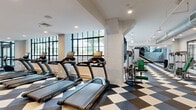 Located at 40 Locke St in Haverhill, MA, this expansive fitness center offers a thoughtfully designed, well-equipped space for a comprehensive workout experience. The facility is organized into distinct zones, each tailored to a specific training style, and is unified by a striking black-and-white checkered tile floor that flows throughout the main areas.

The cardio zone is lined with True-brand treadmills and elliptical machines positioned along floor-to-ceiling steel-framed windows, flooding the space with abundant natural light and offering views of the surrounding greenery. Structural columns finished in white complement the clean, modern aesthetic throughout.

An adjoining strength training area features a variety of cable machines, resistance equipment, and a dedicated free weights section with a full rack of dumbbells and medicine balls. Mirrored walls enhance the sense of openness and allow users to monitor their form throughout their workout.

A standout feature is the functional training room, finished with indoor artificial turf and soaring ceilings. This area is equipped with a multi-functional rig, battle ropes, benches, and a stair-climbing apparatus, making it ideal for high-intensity and functional fitness training. Patterned accent walls and large windows add visual interest and further connect the interior to the outdoors. The facility also includes a small lift for accessibility between levels.