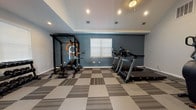 Located at 2109 Oglethorpe Circle in Pooler, Georgia, this dedicated fitness facility is thoughtfully designed to support a comprehensive workout experience. The single-floor space features a vaulted ceiling with recessed lighting that enhances the open, airy feel of the room. The interior is finished with a neutral and blue-gray color palette, lending the space a clean, motivating atmosphere.

The flooring consists of durable, patterned carpet tiles in a checkerboard-style arrangement of gray and beige tones, providing both comfort and practicality underfoot. The strength training area is anchored by a multi-function power rack with a weight bench, complemented by a full dumbbell rack and a selection of kettlebells for free-weight training. Colorful stability and medicine balls are neatly organized on a dedicated rack near a large wall-mounted mirror, which effectively amplifies the sense of space.

The cardio section is well-equipped with multiple treadmills, a stationary air bike, a spin bike, and a standalone cardio stepper, offering diverse options for aerobic exercise. A wall-mounted flat-screen television is positioned for convenient viewing during workouts. Yoga and exercise mats are wall-mounted for easy access. Natural light filters in through multiple windows fitted with white blinds, and a glass-panel door provides direct exterior access, ensuring the space remains bright and well-ventilated.
