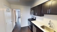 This single-floor space offers a clean and functional layout with thoughtful design details throughout. The kitchen is a standout feature, showcasing rich dark espresso-toned cabinetry that contrasts beautifully against light countertops, providing ample storage and workspace. Stainless steel appliances, including a gas range, built-in microwave, and dishwasher, give the kitchen a modern, polished look. The sink is conveniently positioned along the counter, completing the efficient galley-style layout. Dark hardwood flooring runs seamlessly throughout the space, adding warmth and a sense of continuity from room to room. The neutral, light-toned walls create a bright and open ambiance, allowing natural light to travel through the connecting hallway and into adjoining rooms. The hallway leads further into the residence, hinting at additional living spaces beyond. The overall aesthetic is contemporary and move-in ready, balancing practicality with modern style. Clean lines, consistent finishes, and a neutral color palette make this a versatile space well-suited to a variety of décor preferences.