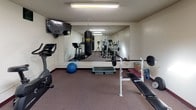 This fitness center is a comprehensively outfitted single-room facility designed to support a wide range of workout routines. The space is carpeted throughout and illuminated by a combination of a central skylight and fluorescent ceiling fixtures, providing consistent, even lighting across the room. A large wall mirror along one side visually expands the room while allowing users to monitor their form during exercise.

Cardio equipment includes a treadmill, stationary bike, and multiple elliptical machines, offering varied options for aerobic training. For strength training, the facility provides a multi-station cable machine, a weight bench with barbell and plates, and a hanging Everlast punching bag for boxing-style workouts. A stability ball and step platform round out the functional fitness options.

A wall-mounted flat-screen television adds an entertainment element to cardio sessions. Signage on the walls indicates facility hours and usage guidelines, reflecting an organized and rules-conscious environment. The neutral-toned walls and dark baseboards give the space a clean, utilitarian appearance suited to focused exercise. Overall, the fitness center delivers a practical and varied workout environment within a compact, efficiently arranged layout.