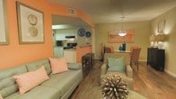 This single-floor space showcases an open-concept layout that seamlessly connects the living, dining, and kitchen areas into one cohesive environment. The interior is distinguished by bold orange accent walls that contrast warmly against neutral beige tones, creating an energetic yet inviting ambiance throughout.

The living area features a comfortable sofa with coordinating accent pillows in coral and teal, complemented by hardwood-style flooring that extends throughout the space. A woven area rug and decorative elements, including a sculptural centerpiece, add texture and visual interest to the room.

The kitchen is accessible via an open pass-through design, featuring white cabinetry, a black range, and a raised counter with bar seating — ideal for casual dining. Decorative wall plates add a personalized touch to the kitchen space.

Adjacent to the living area, the dining space accommodates a round table with upholstered chairs in warm orange tones, illuminated by a stylish pendant light. An abstract artwork in coordinating colors anchors the dining area's wall. The consistent color palette and open flow between zones make this a well-connected and visually cohesive living space.