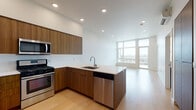 This contemporary single-floor residence showcases a clean, modern design with an open-concept layout that seamlessly connects the kitchen and living areas. The kitchen is a standout feature, equipped with warm walnut-toned cabinetry, a large peninsula with a white quartz countertop and an undermount sink, and a full suite of stainless steel appliances including a gas range, microwave, dishwasher, and refrigerator. Recessed lighting throughout keeps the space bright and well-illuminated.

The living area is bathed in natural light thanks to expansive floor-to-ceiling windows and a glass door that opens onto a balcony, offering sweeping views of the surrounding cityscape. Light hardwood flooring flows consistently throughout the main living spaces, enhancing the airy, open ambiance.

The bathroom maintains the same refined aesthetic, featuring a long walnut vanity with white countertop, an illuminated mirror, light-toned tile flooring, and clean white walls that create a calm, minimalist atmosphere.

A washer and dryer unit is also included within the space, adding practical convenience. Overall, the unit presents a cohesive, contemporary design with quality finishes, generous natural light, and a functional open floor plan.