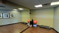 This single-floor fitness studio is designed to support a versatile range of workout activities in a clean, focused environment. The room features durable hardwood-style flooring that provides both aesthetic appeal and practical support for a variety of exercises. A large wall-to-ceiling mirror spans one side of the room, allowing users to monitor their form and making the space feel more open and expansive.

The studio is outfitted with a solid selection of functional fitness equipment, including stability balls, a BOSU balance trainer, colorful kettlebells in varying weights, and aerobic step platforms — catering to strength, balance, and cardio training needs. A foam roller or similar cylindrical equipment is also present for recovery and mobility work.

Overhead, a suspended acoustic tile ceiling houses recessed panel lighting that delivers consistent, even illumination throughout the space. The walls are painted in a soft sage green tone, contributing to a calm and focused atmosphere conducive to exercise. Dark wood base trim adds a finished, polished look to the room's perimeter. A framed piece of wall art adds a subtle decorative touch. Overall, the studio strikes a practical yet inviting balance, making it well-suited for personal training, group fitness classes, or individual workouts.