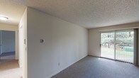 This single-floor unit offers a straightforward, functional layout with clearly defined living spaces. The main living area is generously sized, finished with low-pile gray carpeting and neutral white walls that create a clean, versatile backdrop. A textured popcorn ceiling runs throughout the space, and natural light enters through a sliding glass door that opens to a view of mature trees and greenery. A wall-mounted air conditioning unit and baseboard heating system along the perimeter walls provide year-round climate control.

The kitchen is efficiently arranged and features warm, medium-toned oak cabinetry spanning both upper and lower sections, offering ample storage. Light-colored countertops provide functional workspace, and the kitchen is equipped with a white electric coil range, an overhead range hood, and a stainless steel single-basin sink. Tile flooring completes the kitchen area.

The bathroom is finished with tile flooring and includes an oak vanity with a single sink, a large mirror with overhead lighting, a toilet, and a bathtub with shower. The overall aesthetic of the unit is practical and neutral, well-suited for straightforward, comfortable everyday living.