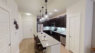 This well-appointed single-floor residence offers a thoughtfully designed open-concept layout that maximizes both functionality and style. The kitchen serves as the centerpiece of the space, showcasing rich dark cabinetry that extends to the ceiling, creating a bold contrast against the crisp white walls and light wood-look flooring that runs throughout. A large granite-topped island anchors the kitchen, providing generous countertop workspace alongside comfortable bar seating for multiple guests — ideal for casual dining or entertaining. Three elegant glass pendant lights hang above the island, adding warmth and visual interest to the space. The kitchen is fully equipped with a complete suite of stainless steel appliances, including a refrigerator, microwave, electric range, and dishwasher. A classic white subway tile backsplash adds a clean, timeless detail behind the cooktop and sink area. Recessed lighting throughout ensures the space is bright and welcoming at all times. A white entry door with modern hardware is visible from the kitchen, contributing to the cohesive, contemporary aesthetic of the home. Additional doors suggest further living spaces beyond the main open area. The overall ambiance is clean, modern, and move-in ready.