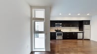 This single-floor space showcases a clean, contemporary design with an open-concept layout that seamlessly connects the living and kitchen areas. The kitchen is a standout feature, outfitted with rich, dark espresso cabinetry that extends to the ceiling, offering ample storage and a bold visual contrast against the bright white walls and light backsplash. Stainless steel appliances, including a gas range, built-in microwave, and a sink set along the window-facing counter, complete the modern culinary space. Recessed ceiling lighting provides even, ambient illumination throughout the kitchen area. Warm, dark hardwood flooring runs continuously throughout the space, adding depth and a sense of cohesion to the open floor plan. Natural light filters in through a double-pane window with horizontal blinds, lending an airy quality to the interior while offering a view to the outside. The crisp white walls and clean architectural lines contribute to a minimalist aesthetic, making the space feel bright, functional, and well-appointed. Overall, the space strikes a refined balance between modern style and practical design.