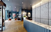 This elegantly designed single-floor space makes a strong first impression with a striking reception area anchored by a fluted navy blue desk topped with a marble surface. Behind it, a large wall of dimensional white relief panels with organic botanical motifs serves as a dramatic focal point, illuminated by warm cove lighting above. A cascading chandelier with layered white fringe adds a refined decorative touch overhead, while wide-plank hardwood flooring flows throughout the entire space.

A comfortable lounge area features full-height steel-blue paneled walls, a plush velvet sofa in warm caramel tones, a substantial marble coffee table, and an ornate area rug. Artistic wall sconces and a floor lamp contribute to the warm, curated ambiance. A separate seating nook offers bold red curved chairs arranged around a sculptural side table, accompanied by floor-to-ceiling windows that invite abundant natural light and outward views.

A built-in beverage station with dark cabinetry, gold hardware, and a marble countertop provides an upscale hospitality touch. Artwork is thoughtfully displayed throughout, enhancing the gallery-like quality of the space. The overall atmosphere is polished, sophisticated, and thoughtfully composed.