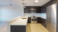 Located at 1548 Hancock Street in Quincy, Massachusetts, this single-floor unit showcases a clean, modern aesthetic throughout. The open-concept living and kitchen area is anchored by light hardwood-style flooring that runs seamlessly across the space, creating a bright and cohesive atmosphere. The kitchen is well-appointed with dark espresso cabinetry, white quartz countertops, a tile backsplash, and stainless steel appliances including a range, microwave, and refrigerator. Pendant lighting above the kitchen island adds a contemporary touch, while the peninsula layout provides a natural transition into the living area.

The living space is open and airy with neutral walls and recessed lighting. The bedroom features plush carpeting, generous natural light from large windows, and urban views of the surrounding Quincy cityscape. A second room with hardwood-style flooring also benefits from oversized windows with city views, offering flexible use as a bedroom or office.

The bathroom is finished in a clean, minimalist style with tile flooring, a walk-in shower, a dark-toned vanity, and an LED-lit mirror, lending a polished, hotel-like quality. Overall, the unit presents a cohesive modern design with quality finishes throughout.