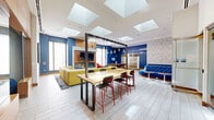 Located at 744 Columbus Ave in Boston's dynamic South End/Roxbury neighborhood, this thoughtfully designed amenity space spans a single expansive floor, offering a rich variety of functional and social environments. The main living area commands attention with soaring ceilings, skylights that flood the space with natural light, and an energetic mix of bold furnishings — including a vibrant yellow sectional sofa, a tufted blue curved banquette, and an organic-shaped sculptural wood coffee table — all set atop patterned area rugs over light hardwood-style tile flooring. A striking large-scale mural celebrating local Boston culture anchors an open entry corridor with clean white walls and warm wood-paneled accents. A cozy media lounge features a richly paneled wood wall, a sculptural red sofa, a large flat-screen TV mounted on a textured tile accent wall, and a diagonal diamond-pattern built-in shelving unit. The business center showcases a bold geometric-tiled feature wall and a row of desktop workstations beneath pendant lighting. A game room with deep blue walls houses a full-size billiards table and glass-partitioned spaces. Private meeting rooms with colorful carpeting and locally themed artwork provide dedicated collaborative spaces, completing this dynamic, community-centered amenity floor.