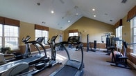 This fitness center offers a thoughtfully designed workout space that accommodates a wide range of exercise needs. The room is defined by its striking vaulted ceiling, which rises to a dramatic peak and is fitted with recessed lighting and a ceiling fan to keep the space bright and well-ventilated. Warm, neutral-toned walls complement the carpeted flooring, creating an inviting and comfortable environment for exercise.

The cardio section is well-appointed with multiple treadmills and elliptical machines, positioned to take advantage of the natural light that streams in through several windows dressed with warm Roman shades. The windows provide a pleasant connection to the outdoors while working out. Toward the rear of the space, a multi-station strength training machine and a cable weight system offer versatile options for resistance and strength exercises, rounding out the facility's capabilities.

The open layout ensures that equipment is generously spaced, allowing users to move freely and safely between stations. The combination of vaulted ceilings and natural light gives the room an airy, expansive feel despite its focused single-floor footprint. Overall, this fitness center is a fully functional and welcoming facility suitable for both cardio and strength training routines.