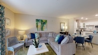 Located at 220 Pine Knot Lane in Graham, North Carolina, this single-floor residence offers a cohesive and well-designed living environment with a calm, coastal-inspired aesthetic throughout. The open-concept layout connects the living, dining, and kitchen areas, creating a sense of generous, flowing space ideal for both everyday living and entertaining.

The living room serves as the heart of the home, featuring neutral walls, plush carpeting, and large windows dressed with floral-patterned drapes that invite natural light while adding a decorative touch. The space comfortably accommodates a large sectional sofa and a cozy sitting area.

The kitchen is outfitted with white shaker-style cabinetry, granite countertops, and a full suite of stainless steel appliances, including a range with a stainless hood, built-in microwave, dishwasher, and refrigerator. Open glass-front upper cabinets add an elegant detail.

Two well-proportioned bedrooms each feature ceiling fans, soft carpet flooring, and ample room for furnishings. Both bathrooms are finished with tile flooring, granite vanities, white cabinetry, and tub-shower combinations with built-in shelving, maintaining a clean and consistent design throughout the residence.