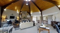The clubroom is an inviting and architecturally distinctive gathering space defined by its soaring vaulted ceiling clad in warm wood planking and supported by bold arched dark-wood beams that converge at a central peak. This striking overhead feature immediately draws the eye upward, creating an expansive, lodge-like atmosphere throughout the room.

At the heart of the space stands a full brick fireplace, serving as a natural focal point and adding a rustic, cozy character to the room. Surrounding it, comfortable lounge seating — including a cream sofa, leather armchairs, and accent tables — creates a relaxed social area ideal for gatherings or quiet downtime.

Along one wall, a dedicated workstation area features two computer desks, office chairs, and a wall-mounted flat-screen television flanked by decorative accents, offering a functional space for residents to work or connect.

Large glass French doors flood the room with natural light while providing direct views and access to the outdoor surroundings. Brick accent walls and columns echo the fireplace's rustic detailing throughout. An adjacent area with bar-height seating and a pass-through counter adds to the room's versatility, making the clubroom well-suited for both everyday use and community events.