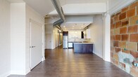 Located at 230 Scott St in Buffalo, NY, this single-floor loft unit showcases a striking blend of industrial character and modern updates. The spacious open main area features exposed brick walls, soaring open-beam ceilings with visible ductwork, and a skylight that draws natural light deep into the space. Flooring transitions between durable luxury vinyl planks and patterned carpet, defining distinct zones within the open layout. A sleek kitchen is outfitted with warm wood cabinetry, stainless steel appliances including a refrigerator, and under-cabinet lighting, offering a functional and stylish cooking area. A steel-railed staircase with a modern horizontal balustrade adds an architectural focal point and connects levels within the building. The bathroom is finished with light tile flooring, a granite-topped vanity, and a walk-in shower. A dedicated laundry room provides added convenience with a full-size front-loading washer and dryer, shelving, and an on-site water heater. Throughout the unit, crisp white walls contrast with the raw industrial elements, creating an ambiance that is both contemporary and full of character — well-suited for a variety of uses.