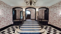 This distinguished residential lobby makes a powerful first impression through its carefully preserved classical design. The centerpiece of the space is a sweeping marble staircase with dark wrought iron railings, its broad steps curving gracefully upward from a dramatic rounded base. The flooring throughout the entry level features a bold black-and-white checkered marble pattern that reinforces the space's formal, stately character.

Every wall is adorned with intricate red-and-white toile wallpaper, depicting detailed pastoral and figurative scenes that lend the space a refined, historic elegance. Dark, arched doorframes provide strong architectural contrast against the lighter wallpaper, while ornate brass wall sconces cast a warm, ambient glow throughout. A decorative chandelier with candelabra-style lighting hangs overhead, completing the classic aesthetic.

A gilt-framed mirror rests above a marble console table along one wall, adding depth and sophistication to the lobby. Resident mailboxes are neatly integrated along an adjacent wall, providing practical functionality without disrupting the overall elegance. The upper stairwell continues the toile wallpaper treatment alongside carpeted treads and black-trimmed banisters, maintaining a cohesive design language throughout. The space balances timeless grandeur with the functional needs of a residential building.