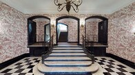This distinguished residential lobby makes a powerful first impression through its carefully preserved classical design. The centerpiece of the space is a sweeping marble staircase with dark wrought iron railings, its broad steps curving gracefully upward from a dramatic rounded base. The flooring throughout the entry level features a bold black-and-white checkered marble pattern that reinforces the space's formal, stately character.

Every wall is adorned with intricate red-and-white toile wallpaper, depicting detailed pastoral and figurative scenes that lend the space a refined, historic elegance. Dark, arched doorframes provide strong architectural contrast against the lighter wallpaper, while ornate brass wall sconces cast a warm, ambient glow throughout. A decorative chandelier with candelabra-style lighting hangs overhead, completing the classic aesthetic.

A gilt-framed mirror rests above a marble console table along one wall, adding depth and sophistication to the lobby. Resident mailboxes are neatly integrated along an adjacent wall, providing practical functionality without disrupting the overall elegance. The upper stairwell continues the toile wallpaper treatment alongside carpeted treads and black-trimmed banisters, maintaining a cohesive design language throughout. The space balances timeless grandeur with the functional needs of a residential building.