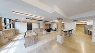 This single-floor space masterfully combines historic architectural elements with contemporary finishes, creating a distinctive and functional environment. Polished concrete floors extend throughout, reflecting light and lending a clean, open quality to the interior. Substantial reclaimed timber columns and diagonal braces anchor the space structurally while serving as striking design features that highlight the building's industrial heritage.

The main open area accommodates multiple collaborative zones, including a long wooden conference table with seating for several occupants and a raised bar-height table with stools suited for informal gatherings. Exposed copper piping and industrial-style linear lighting fixtures run along the ceiling, complementing the raw aesthetic. A dedicated lounge area centers around a modern linear fireplace set against a textured reclaimed wood accent wall, flanked by navy upholstered armchairs for a refined yet relaxed setting.

Natural light enters through black-framed multi-pane windows, offering views of surrounding greenery. A fully equipped kitchenette features stone countertops, a stainless steel sink with a professional-style faucet, and refrigeration. Additional private rooms accessible via glass-paned doors complete the layout, providing flexibility for a variety of professional or event uses.