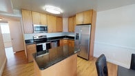 This single-floor residence offers a thoughtfully designed layout that balances functionality and comfort. The kitchen is a standout feature, showcasing rich dark granite countertops atop a central island, warm maple cabinetry, and a full suite of stainless steel appliances including a French door refrigerator with water and ice dispenser, a smooth-top electric range with built-in microwave, and a dishwasher. Light hardwood flooring runs throughout the kitchen, adding warmth to the space. The layout is efficiently arranged in an L-shape with the island providing additional prep and seating space. The living area offers a welcoming atmosphere with neutral-toned carpet, clean white walls, and tasteful, understated décor. A plush area rug anchors the seating arrangement, which centers around a low-profile coffee table. The room is softly lit and maintains a calm, relaxed ambiance. Together, these spaces create a cohesive and livable environment that is both practical and inviting, well-suited for everyday living and casual entertaining.