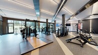 Located at 630 Washington Ave in Belleville, NJ, this premium fitness center offers a generously sized, single-floor exercise facility designed with both functionality and aesthetics in mind. The space is defined by its contemporary architecture, featuring high ceilings with bold linear LED lighting that runs throughout, dark cylindrical columns, and a sophisticated color palette of charcoal, white, and warm wood tones.

The main workout floor is thoughtfully zoned to accommodate a wide range of fitness activities. A comprehensive selection of strength training equipment — including cable machines, multi-functional rigs, benches, and free weights — is arranged across rubber flooring with inlaid contrast striping that guides movement through the space. A row of treadmills and cardio machines lines a full-height glass curtain wall, offering unobstructed views of the surrounding greenery and abundant natural light.

A dedicated yoga and functional training studio, enclosed by sleek floor-to-ceiling glass partitions, provides a more intimate space for focused workouts. Inside, a wall-mounted flat-screen TV, a heavy bag, suspension trainers, and a stylized marbled accent wall create a dynamic yet serene environment. Additional flat-screen displays are positioned throughout the main floor to keep users engaged. The overall ambiance is that of a high-end, boutique fitness facility built to support a full spectrum of training needs.
