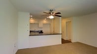 This single-floor residence is designed with an open-concept layout that seamlessly connects the kitchen and living areas, creating a spacious and functional environment. The kitchen is defined by a raised peninsula with dark granite countertops that provide ample prep and serving space while acting as a natural divider between the cooking and living zones. White upper cabinetry with recessed panel doors lines the kitchen walls, offering generous storage, while recessed ceiling lights illuminate the workspace effectively. A stainless steel sink is positioned along the back wall of the kitchen, adding a clean, modern touch. The living area features warm, neutral-toned carpeting and is centered beneath a classic ceiling fan with an integrated light fixture, ensuring comfortable airflow throughout the space. A textured ceiling adds a traditional finish overhead. Neutral, light-colored walls throughout the space contribute to a bright and airy atmosphere. A doorway visible from the main living area leads to an additional room or hallway, suggesting a well-connected floor plan. The transition from carpet in the living area to hard flooring in the adjoining space adds practical variety to the overall design.