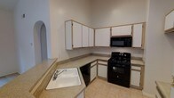 This single-floor residence offers a practical and well-organized layout designed for comfortable everyday living. The kitchen is a standout feature, showcasing white cabinetry with light wood trim that extends to the ceiling, maximizing storage space throughout. A suite of black appliances — including a gas range, built-in microwave, and dishwasher — provides a bold contrast against the lighter cabinetry and countertops. The counters offer generous prep space, complemented by a white drop-in sink conveniently positioned on a peninsula that helps define the kitchen area. Light neutral tile flooring runs underfoot, adding to the clean and cohesive aesthetic. A distinctive arched entryway is visible from the kitchen, lending an elegant architectural touch that hints at a thoughtfully designed floor plan throughout the home. The vaulted ceiling height in the kitchen creates an open, airy atmosphere, adding to the overall sense of space. The neutral color palette throughout provides a versatile backdrop, making the home move-in ready and easy to personalize.