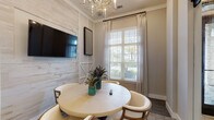 This conference room in Fernandina Beach, Florida, is part of a larger, thoughtfully designed facility that blends coastal sophistication with functional meeting space. The conference room itself centers around a round light-wood table surrounded by cushioned, wood-framed chairs, creating an intimate and collaborative atmosphere ideal for small meetings or consultations.

A wall-mounted flat-screen television is positioned on a textured stone accent wall, accompanied by decorative wall paneling featuring geometric cross-pattern millwork details. An ornate, branch-style chandelier overhead adds an elegant focal point, while crown molding reinforces the refined aesthetic throughout. A large window dressed with sheer linen curtains and white blinds allows generous natural light to filter in, offering views of the surrounding landscape.

The broader facility visible from the conference room reveals soaring ceilings with exposed wood beams, recessed lighting, light-toned tile flooring, and structured white columns that define open gathering areas. A glass-paneled entryway with dark-framed doors provides a grand, welcoming entrance to the building. The overall ambiance is warm, polished, and well-appointed, reflecting a carefully curated coastal design sensibility throughout.