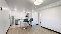 This efficiently designed studio unit offers a clean, modern aesthetic throughout its single floor, making smart use of every square foot. The open-concept layout connects the living and kitchen areas seamlessly, anchored by light wood-look luxury vinyl plank flooring and crisp white walls that create a bright, airy atmosphere.

The kitchen is well-appointed with a full suite of stainless steel appliances, including a full-size refrigerator, dishwasher, electric range with overhead ventilation, and a built-in microwave. White cabinetry with sleek hardware provides ample storage, while the clean white countertops extend generously along the walls. A freestanding white bar-height table paired with two black adjustable bar stools serves as a casual dining and gathering spot just off the kitchen.

The bedroom area features a large wood-panel headboard wall with integrated side shelving and is accented by soft blue tones that lend a calming character to the space. A window with outdoor balcony access fills the room with natural light. A separate entryway with a secure door and window provides a defined transition into the unit.

The bathroom is finished in a cool blue palette, featuring a walk-in shower with white textured tile surround, chrome fixtures, and a frosted glass door — clean and functional in design.