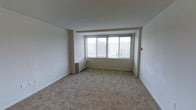 This single-floor space features a generously sized main living area finished with wall-to-wall carpeting in a neutral beige tone, contributing to a warm and understated aesthetic. The room is characterized by clean white walls and a simple, unadorned ceiling that lends a bright, open feel to the interior.

A large, multi-pane picture window serves as a central architectural feature of the living area, flooding the space with natural light while offering elevated views of the surrounding urban cityscape. The window is framed by a recessed surround, adding subtle depth to the wall composition.

Heating is provided by a wall-mounted radiator unit positioned beneath the window, a practical and space-efficient solution commonly found in urban multi-unit buildings. The overall color palette remains consistently neutral — white walls paired with light carpeting — creating a blank canvas that adapts well to a variety of furnishing styles.

The space maintains a straightforward, functional layout with no unnecessary architectural clutter, making it highly versatile. The elevated vantage point suggested by the outdoor views indicates the unit is situated on an upper floor of a larger building.