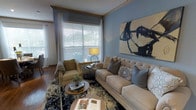 This single-floor space centers around a thoughtfully designed living room that combines comfort and contemporary style. The room is finished with soft blue walls that create a calm, welcoming atmosphere, complemented by warm hardwood flooring that adds a touch of natural elegance. A large window dressed with white horizontal blinds and sheer curtains allows generous natural light to fill the space, offering a view of the greenery outside.

The living room is furnished with a tufted beige sofa adorned with an assortment of patterned and solid pillows in muted blue and grey tones, alongside a classic plaid armchair that adds a traditional touch. A round wooden coffee table anchors the seating arrangement, topped with books and a small potted plant. An ornate area rug with deep blue and ivory tones ties the furniture grouping together cohesively.

A striking abstract artwork in black and neutral tones serves as a bold focal point on the main wall, adding artistic character to the room. Warm ambient lighting from a table lamp with a round metallic base further enhances the inviting atmosphere of this well-curated living space.