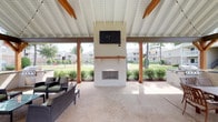 The Grilling Pavilion is a well-appointed outdoor entertaining space designed to provide a comfortable and functional environment for outdoor gatherings. The structure features a covered roof supported by warm wood-toned posts and beams, creating a defined yet open-air atmosphere that allows natural light and breezes to flow freely throughout.

A central white brick fireplace serves as the focal point of the pavilion, extending from floor to ceiling and topped with a wall-mounted flat-screen television — making it equally suited for relaxed social gatherings or outdoor viewing events. Adjacent to the fireplace, a seating area is furnished with rattan-style chairs, offering a comfortable space to gather around the fire.

On the opposite end of the pavilion, a built-in grill station with a countertop surface provides a dedicated cooking area, ideal for outdoor grilling. A pergola-style extension connects seamlessly to the main covered structure, adding architectural interest and framing views of the surrounding landscaped grounds. Colorful flowering shrubs, mature trees, and manicured lawns are visible from all sides, enhancing the open, park-like ambiance of the space. The aggregate concrete flooring is durable and visually cohesive throughout.