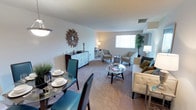 This single-floor unit offers a seamlessly connected living and dining space designed for comfortable, everyday living. The open-plan layout allows the dining and living areas to flow naturally into one another, creating a sense of spaciousness without visual barriers.

The dining area is positioned near the entrance and is furnished with a glass-top table paired with bold teal upholstered chairs, adding a pop of color to the neutral palette. A decorative sunburst mirror and a patterned credenza adorn the adjacent wall, contributing character to the space.

The living area is generously sized, featuring wall-to-wall carpeting in a warm neutral tone that adds softness underfoot. A cream tufted sofa, complementary accent chairs in a subtle geometric pattern, and a round glass coffee table anchor the seating arrangement. A large window allows natural light to fill the room, enhancing the bright, airy atmosphere.

Neutral white walls throughout provide a clean, versatile backdrop, while layered lighting — including floor lamps and an overhead fixture — ensures a well-lit environment at any time of day. A tall potted plant near the window introduces a natural element, rounding out the inviting ambiance of this well-appointed space.