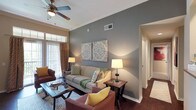 This 2-bedroom, 2-bathroom single-story residence offers a thoughtfully designed layout that maximizes both comfort and functionality. The living room serves as the centerpiece of the home, featuring crown molding that adds an elegant architectural detail along the ceiling perimeter. A ceiling fan provides year-round comfort, while natural light filters in through sliding glass doors dressed with vertical blinds and patterned drape panels, suggesting access to an outdoor area. The living space is anchored by a bold accent wall in a cool grey tone, providing a striking contrast to the warmer neutral tones found throughout the room. The flooring features rich, dark hardwood, adding warmth and depth to the space. The layout flows naturally toward additional rooms via a hallway visible from the main living area, connecting the bedrooms and bathrooms. The overall ambiance is warm and inviting, with layered lighting from floor and table lamps complementing the natural light. Crown molding and carefully considered architectural details elevate the space, creating a polished and cohesive aesthetic throughout this well-proportioned home.